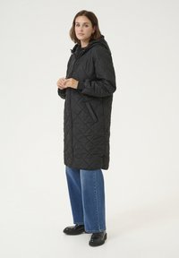 Manteau long en cuir noir matelassé avec capuche, doté d'une fermeture éclair à l'avant, de poches latérales et de poignets élastiques, associé à un jean bleu à jambes larges et des chaussures noires.