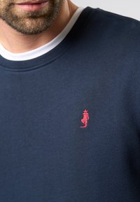 Pulover bleumarin cu guler alb, având un logo roșu brodat al unei siluete pe piept. Textură netedă, design casual.