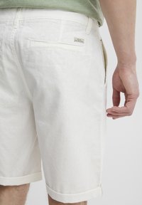 Pantaloni corti bianchi in lino con risvolti, dotati di una tasca sul retro e di un piccolo logo sul lato destro. Tessuto morbido e leggero.