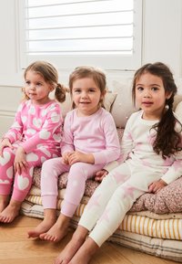 Roosad pidžaamad suurte valgete südametega, valged pidžaamad roosade tähtedega, kõik pehmest kangast, kantud kolme lapse poolt, kes istuvad diivanil.