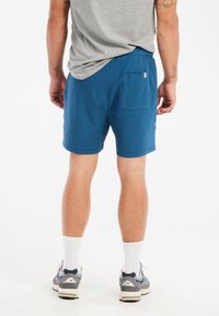Blå træningsshorts med struktureret stof, elastisk talje og en baglomme. Bæres med en grå t-shirt og sneakers.