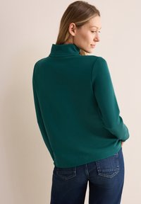 Teal Pullover mit hohem Kragen, aus weichem Stoff gefertigt. Verfügt über lange Ärmel und einen lässigen Schnitt, kombiniert mit dunklen Jeans.