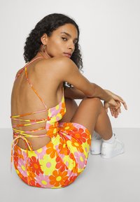 Vestido floral colorido com padrões em rosa, laranja e amarelo. As costas estão parcialmente expostas, com alças cruzadas e um detalhe de laço lateral.