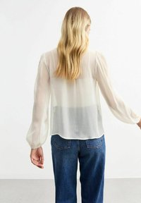 Sheer white blouse with long, puffed sleeves and a pleated texture, paired with blue jeans. Light fabric, relaxed fit, soft drape.