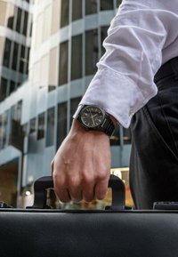 Montre analogique noire sur un poignet avec un poignet de chemise blanche, tenant la poignée d'une mallette noire texturée. L'arrière-plan présente des bâtiments en verre.