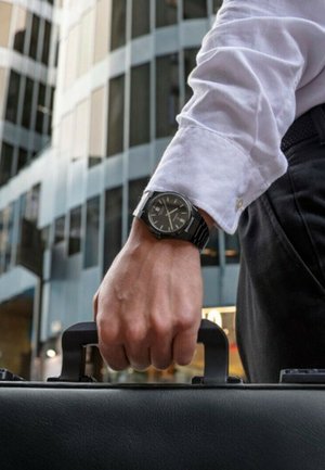 Montre analogique noire sur un poignet avec un poignet de chemise blanche, tenant la poignée d'une mallette noire texturée. L'arrière-plan présente des bâtiments en verre.