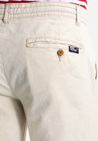 Pantalones beige con un bolsillo trasero con botón y una etiqueta azul navy que dice "Wear The Code", llevados con una camisa de rayas rojas y blancas.