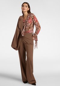 Pantaloni marroni su misura abbinati a una blusa trasparente con motivo floreale nei toni del rosa e del marrone, completati da un blazer marrone.