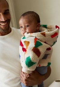 Veste à capuche en polaire multicolore avec des motifs géométriques rouges, verts, orange et marron, portée par un enfant. Texture douce, poignets côtelés.
