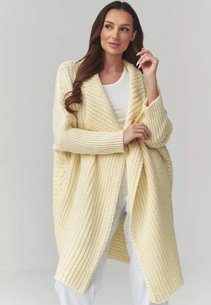 Kobieta o długich brązowych włosach, ubrana w kremowy oversize'owy kardigan z dzianiny, na biały top i spodnie, pozująca z ręką przy twarzy na szarym tle.