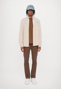 Juostuotas smėlio spalvos marškinėliai su mygtukais, rudas marškinėlis, rudos kelnės, baltos sportinės avalynės ir šviesiai mėlynas skrybėlė su logotipu. Kasdienis apranga.