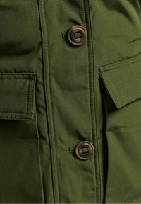 Olijfgroene jas met een gladde textuur, twee klepzakken en grote bruine knopen voor het sluiten.