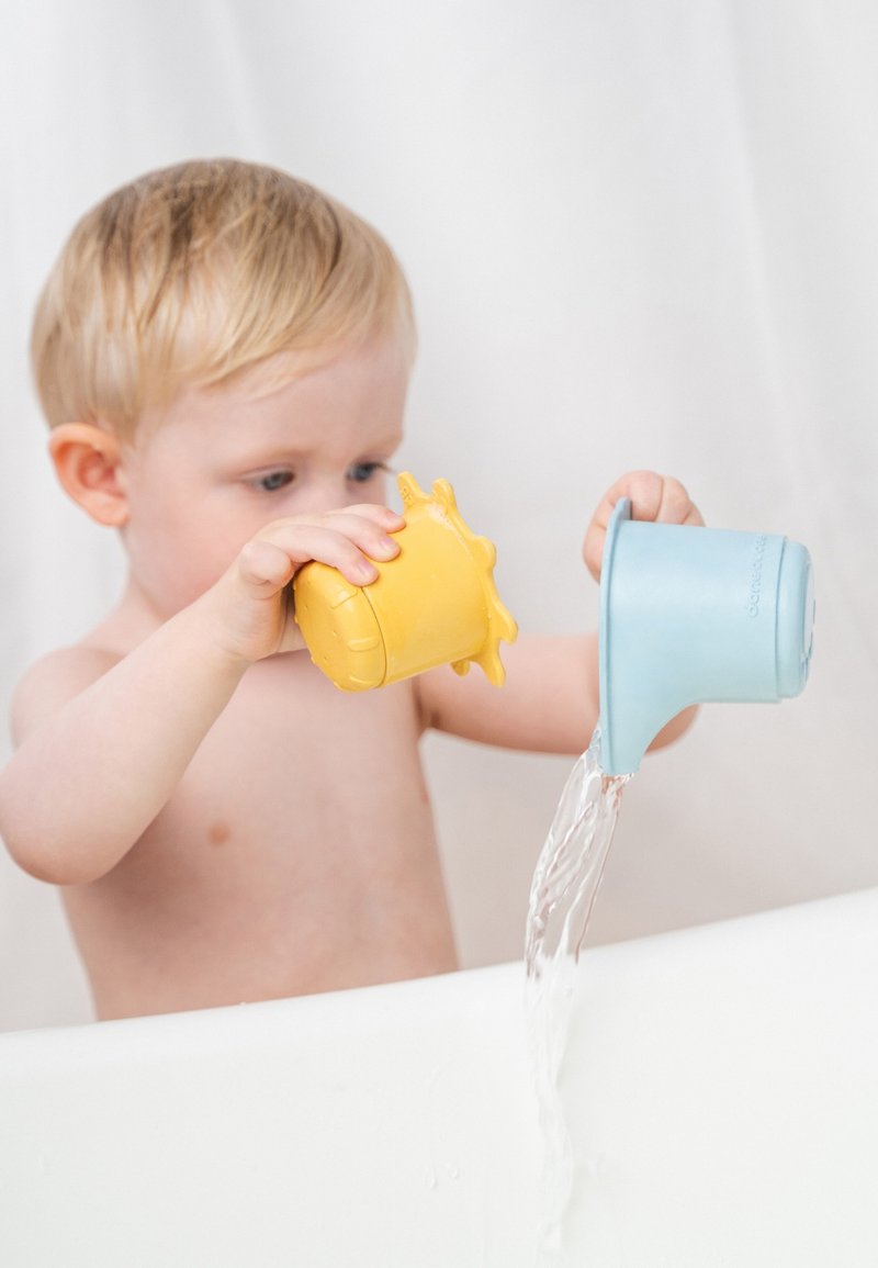 Kind houdt een geel, zonvormig beker in de ene hand en een blauwe beker in de andere, giet water in een badkuip met een witte achtergrond.