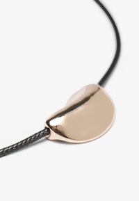Colier din sfoară împletită neagră, cu un pandantiv din metal lucios de culoare aurie, având o formă organică, curbată și o suprafață netedă.