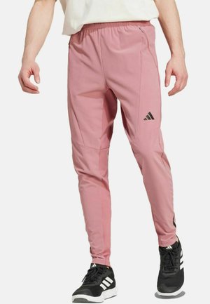 Pink athletic pants with a tapered design, elastic waistband, zipper pockets, and black Adidas logo accents. Worn with black sneakers.