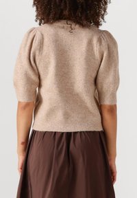 Beige gebreide trui met korte pofmouwen, textuurafwerking en geribde manchetten. Gedragen over een bruine plissérok.