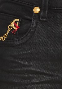 Pantalones de mezclilla negros con un botón dorado y una cadena dorada con un acento de letra. Incluye un detalle de hilo rojo en la cadena.