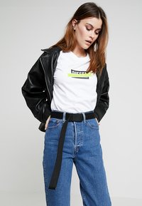 Schwarze Lederjacke über einem weißen T-Shirt mit grünem und schwarzem Logo, kombiniert mit hochgeschnittenen blauen Jeans und einem schwarzen Gürtel.