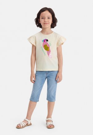 Ung pige med kort mørkt hår iført en cremefarvet kortærmet top med et farverigt papegøjemotiv, lyseblå knælange jeans og hvide sandaler.