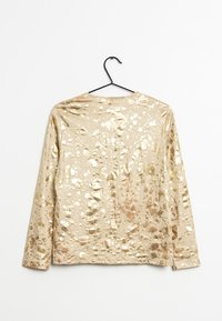 Långärmad guldtop med en glänsande, texturerad yta som har oregelbundna prickiga mönster. Hänger på en svart klädhängare.