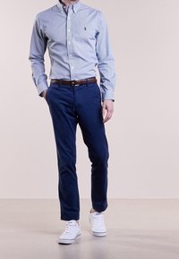 Man som bär en blå randig skjorta med knappar, marinblå byxor, brunt läderbälte och vita sneakers, som går på ett ljust golv.