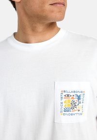 Bijela pamučna majica s ušivenim džepom na prsima, koja sadrži šareni grafički print, uključujući "Billabong" i apstraktne oblike.
