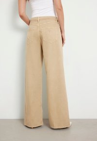 Pantalones de pierna ancha en color beige, confeccionados en tejido suave, con cierre de botón y cremallera, bolsillos traseros y un pequeño logo en el bolsillo derecho.