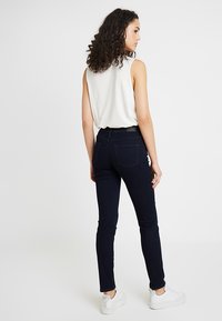 Jeans navy de corte slim con una textura suave, que presentan un clásico diseño de cinco bolsillos y costuras sutiles. Combinados con una blusa sin mangas blanca y zapatos blancos.