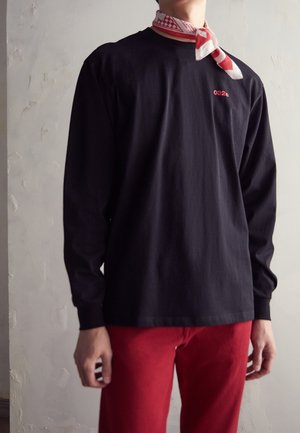 Svart långärmad skjorta med röd broderad "032". Modellen bär röda byxor och en vit scarf med röda rutiga mönster.