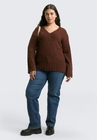 Brązowy sweter z dzianiny z dekoltem w serek, prążkowanymi mankietami i obszyciem, w połączeniu z niebieskimi dżinsami i czarnymi butami na platformie. Modelka trzyma beżową torebkę.