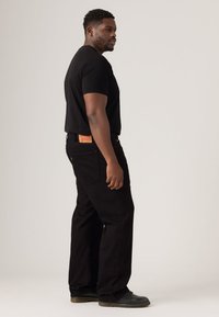 Black short-sleeved t-shirt paired with black pants. Pants feature a numbered tag on the back and a loose fit. Shoes are dark with a thick sole.
