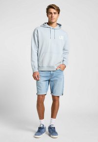 Felpa blu chiaro con logo "LEE" bianco, tasca frontale, abbinata a pantaloni corti di jeans arrotolati e sneakers blu. Calzini bianchi completano l'outfit.