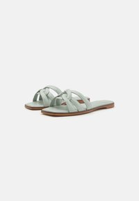 Sandales à glisser en cuir vert clair avec un design de lanière nouée, semelle plate marron et texture lisse. Adaptées pour une tenue décontractée.