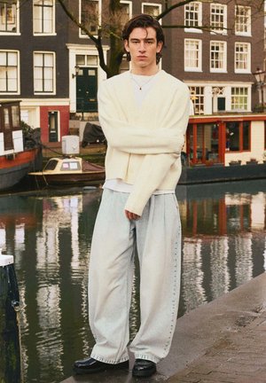 Young man in loose light denim pants, cream cardigan, and black shoes standing by canal with boats and historic buildings in background.