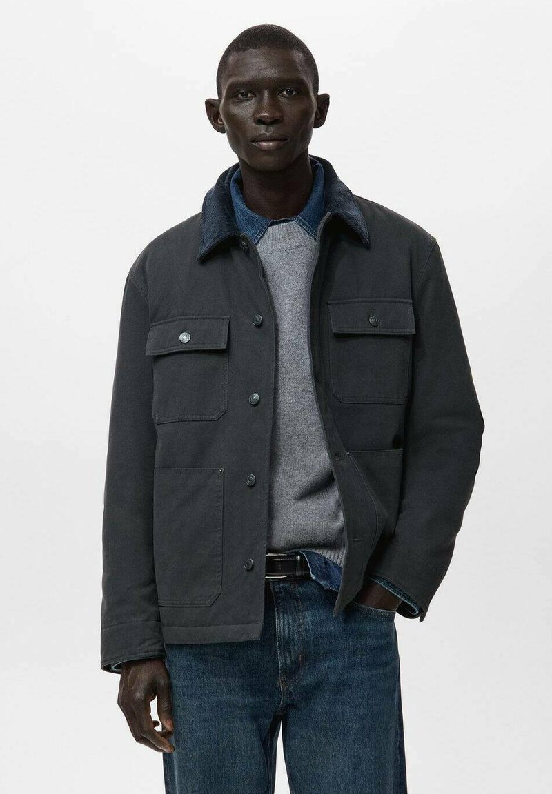 Donkergrijze katoenen jas met een denim kraag, voorzien van twee borstzakken en een knoopsluiting aan de voorkant, gedragen over een grijze trui en jeans.