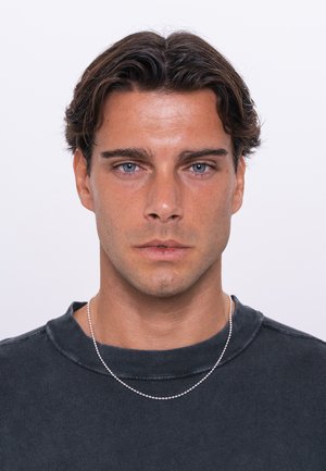 Mladý muž s krátkymi vlnitými hnedými vlasmi a modrými očami, oblečený v tmavom tričku s okrúhlym výstrihom a tenkým strieborným retiazkovým náhrdelníkom, na jednoduchom pozadí.