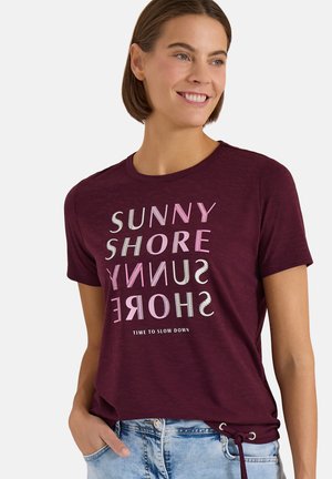 Femme souriante, portant un t-shirt bordeaux avec le texte en miroir "SUNNY SHORE" et la devise "TIME TO SLOW DOWN", associé à un jean bleu clair.