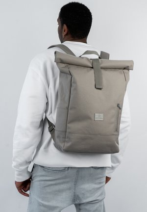 Rucsac gri cu flap, dotat cu un buzunar lateral cu fermoar, curele ajustabile și un material texturat. Design simplu, cu un eticheta pe față.
