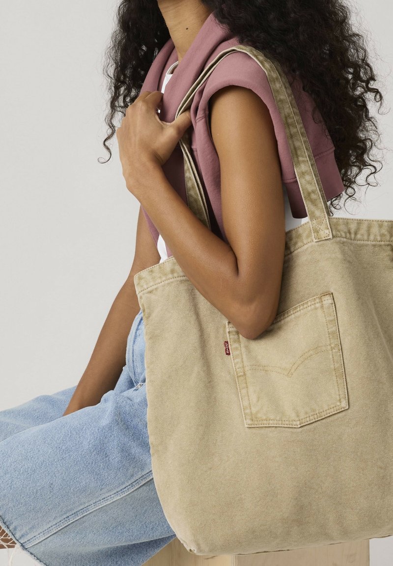 Femme portant un haut sans manches et une jupe en jean, tenant un grand sac cabas en toile beige avec une poche avant sur l'épaule.