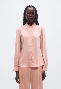 Lyserød satinbluse med mandarin-krave, knaplukning foran og let opsmøgede ærmer. Har subtil tekstur og et lille logodetalje ved kanten.