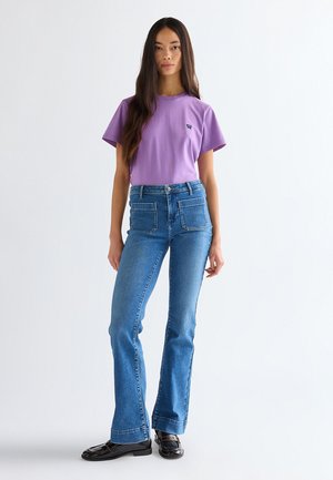 Γυναίκα με μακριά σκοτεινά μαλλιά, που φοράει μωβ μπλουζάκι, τζιν με ψηλή μέση και εμπρός τσέπες, καθώς και μαύρα loafers, στέκεται μπροστά σε λευκό φόντο.