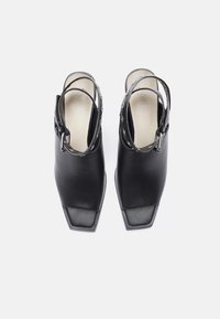 Pari mustien nahkakenkien, joissa on neliön muotoinen kärki ja hopeiset nauhakujat nilkkaremmissä, nähtynä ylhäältä valkoista taustaa vasten.