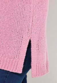 Różowy sweter dziergany o luźnym splotem, z bocznymi rozcięciami i prążkowanymi brzegami. Materiał ma miękką fakturę i swobodny krój, zestawiony z ciemnymi dżinsami.
