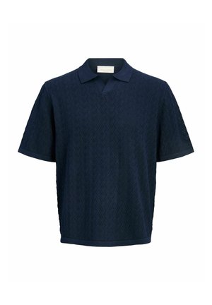 Teenager med krøllet hår iført en mørkeblå kortærmet poloshirt, beige bukser og hvide sneakers, der står og smiler.