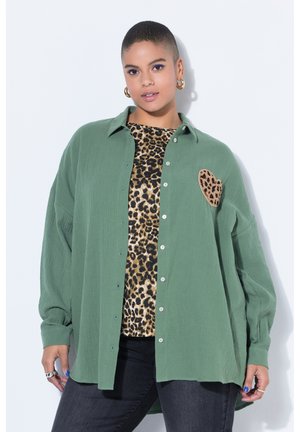 Junge Person mit kurzen Haaren trägt ein grünes, geknöpftes Hemd über einem Leopardenmuster-Top, kombiniert mit dunkler Hose und goldenen Creolen.
