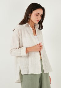 Vaaleanbeige-nappi kauluspaita, joka on valmistettu teksturoidusta kankaasta. Paidassa on pitkät hihat, terävä kaulus ja sivuhalkiot. Vaatetta käytetään valkoisen paidan päällä.