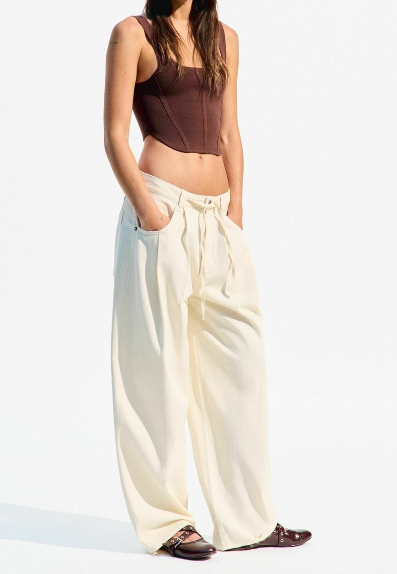 Brown cropped bodysuit, white wide-leg pants with drawstring, and brown shoes with silver accents, against a white background.