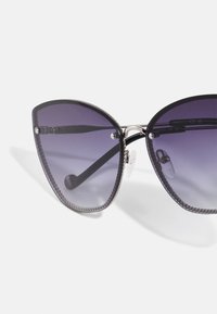 Gafas de sol estilo gato con lentes morados degradados, montura negra, bordes texturizados y acentos metálicos en las patillas, con almohadillas nasales ajustables.