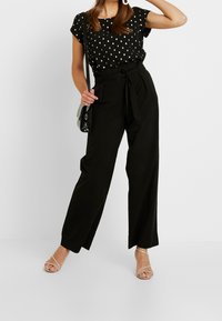 Blusa nera con pois e maniche corte abbinata a pantaloni neri a vita alta. I pantaloni presentano una cintura in tessuto e un design a gamba larga.