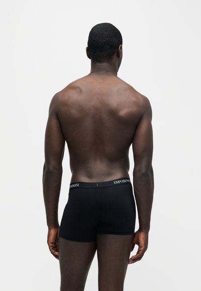 Homme à la peau foncée, dos musclé, portant un boxer noir Emporio Armani, debout devant un fond clair uni, dos à la caméra.